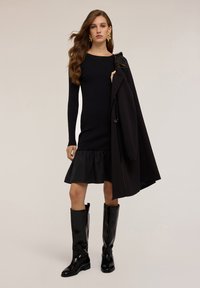 Abito nero a maglia con maniche lunghe e orlo arricciato, abbinato a stivali neri lucidi fino al ginocchio e a un cappotto nero appeso al braccio.