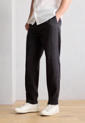 Férfi fekete alkalmi nadrágban és fehér tornacipőben, fakereten állva, egyik kezével a zsebében, sima fal előtt.