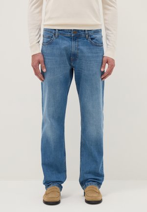Uomo che indossa jeans azzurri a gamba dritta, mocassini beige e una camicia a maniche lunghe color crema, in piedi davanti a uno sfondo neutro.