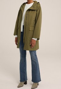 Olive green waterproof parka with a hood, front zipper, two front pockets, and silver hardware; paired with light blue flared jeans and gray boots.