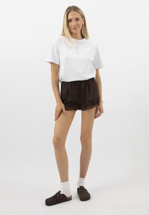 Blonde vrouw staat, draagt een wit t-shirt, zwarte korte broek met kantafwerking, witte sokken en donkere instapschoenen tegen een effen achtergrond.