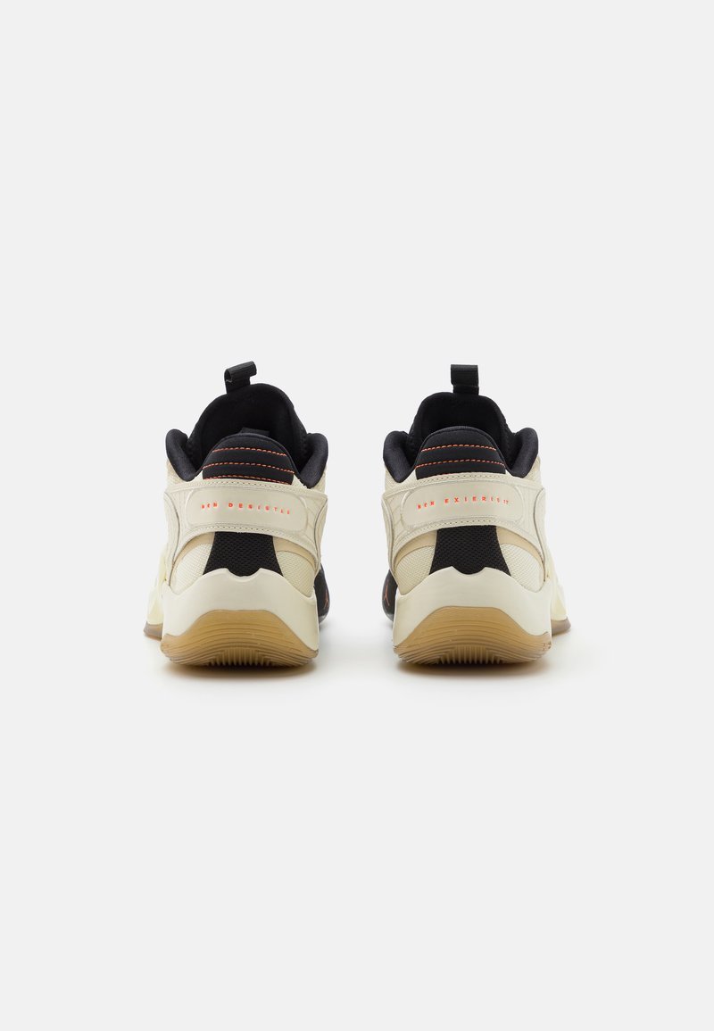 Zapatillas de deporte en crema y negro con un upper texturizado, cuello acolchado y suela de goma con una lengüeta. Acabados rojos en el talón.