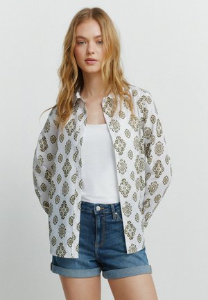 Blonde Frau trägt weißes Tanktop, weißes gemustertes Hemd zum Knöpfen und hochgekrempelte Jeansshorts, steht vor einfachem hellem Hintergrund.