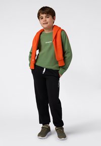 Grønn langermet skjorte med "Champion"-logo, oransje hettegenser drapert over skuldrene, sorte sweatpants og grågrønne joggesko.