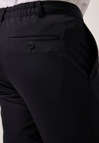 Zwarte trousers van een gladde, rekbare stof met een elastische tailleband en een enkele achterzak die met een knoop is beveiligd.