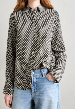 Frau trägt ein schwarz-weißes Hahnentritt-Knopfshemd, das in hellblaue Jeans gesteckt ist, mit einer Hand in der Tasche vor hellem Hintergrund.