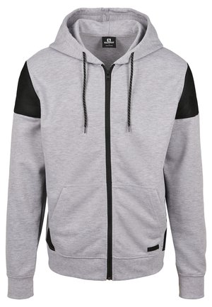 Sweat à capuche gris clair entièrement zippé, avec des accents en mesh noir aux épaules, des poches frontales, une fermeture éclair noire et des cordons noirs à pois blancs.