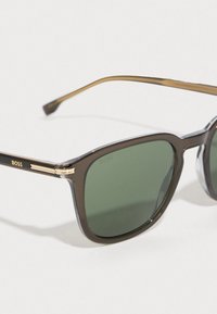 Ορθογώνια γυαλιά ηλίου με σκούρους πράσινους φακούς, μαύρο πλαστικό σκελετό, χρυσές λεπτομέρειες στους βραχίονες και μαλακή υφή. Λογότυπο "BOSS" στο πλάι.