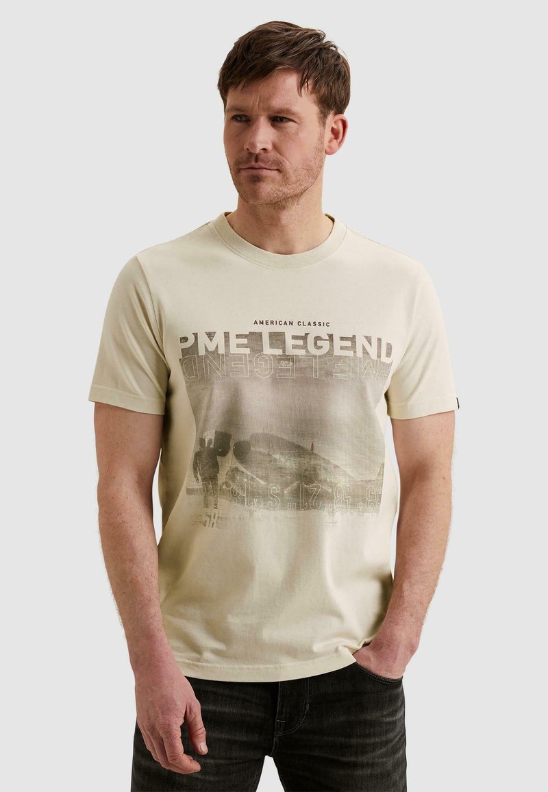 Homme aux cheveux courts portant un t-shirt graphique PME Legend beige clair et un jean foncé, debout avec une main dans la poche.
