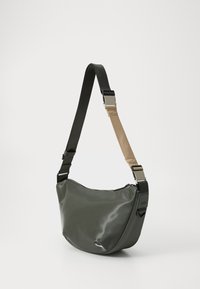 Piccola borsa a tracolla verde con cinturini regolabili neri e beige e chiusura con zip, esposta su uno sfondo bianco.