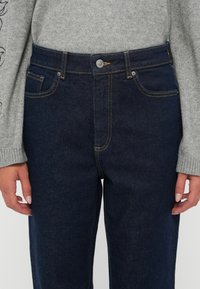 Os jeans de ganga de cor azul escuro apresentam uma cintura alta, bolsos frontais e costuras em contraste. O modelo veste um suéter cinza claro com mangas longas.