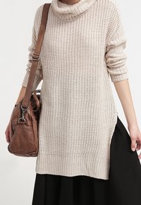 Kobieta w beżowym swetrze z dzianiny z wysokim kołnierzem i czarnej spódnicy, nosząca brązową skórzaną torbę na ramieniu po lewej stronie.