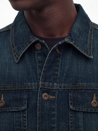 Nahaufnahme einer Person, die eine dunkelblaue Jeansjacke mit braunen Knöpfen und sichtbaren orangefarbenen Nähten über einem schwarzen Hemd trägt.