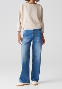 Haut beige texturé avec motif ondulé, manches larges ; jean bleu clair à jambes droites, légèrement délavé ; baskets blanches à semelles plates.