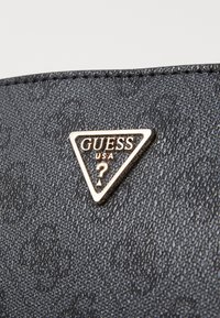Zwart textielmateriaal met een driehoekig gouden logo. Het logo bevat "GUESS" en "USA" in een vet lettertype, met een vraagteken.