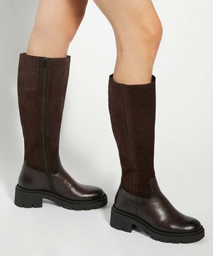 Jambes portant des bottes montantes en cuir et daim marron avec des semelles épaisses noires, présentées sur un fond clair uni.