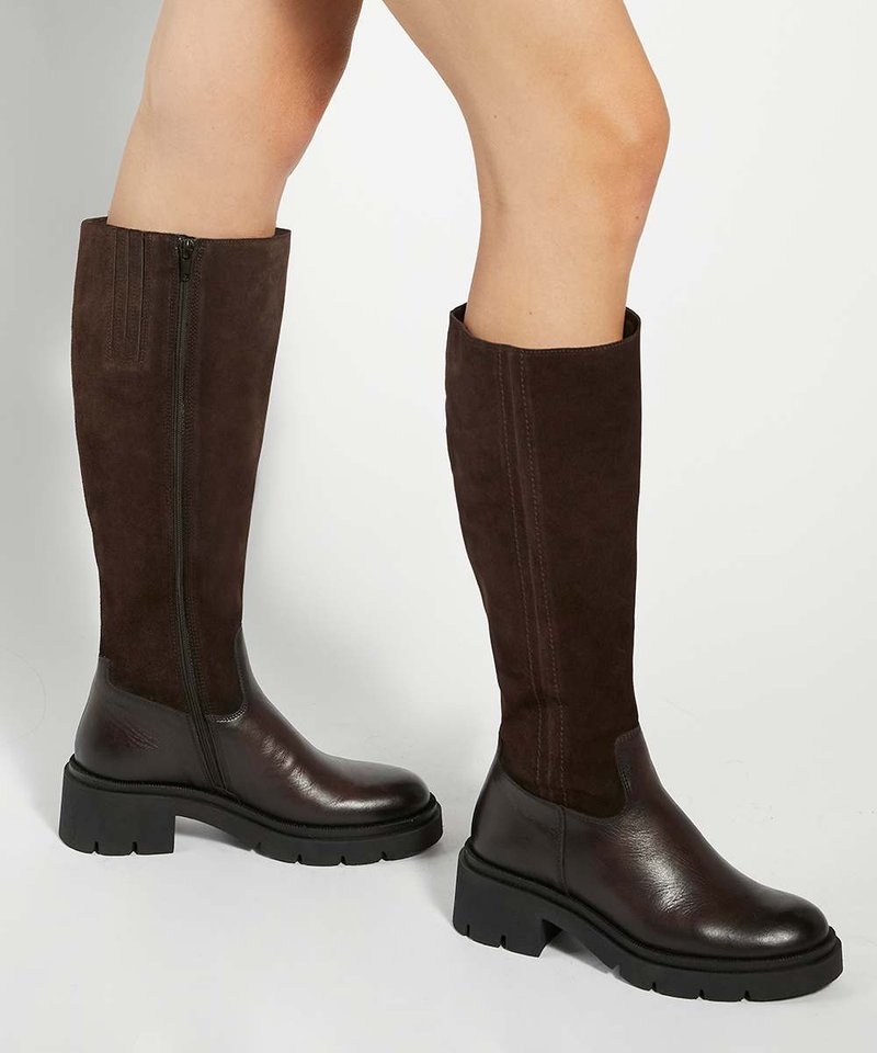 Jambes portant des bottes montantes en cuir et daim marron avec des semelles épaisses noires, présentées sur un fond clair uni.