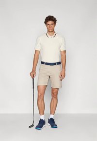 Μπεζ πόλο μπλούζα με γιακά, σορτς σε μπεζ χρώμα, σκούρα μπλε ζώνη και μπλε αθλητικά παπούτσια, κρατώντας ένα μπαστούνι γκολφ, μινιμαλιστικός σχεδιασμός με κοντά μανίκια.