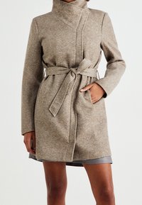 Beigen värinen villasekoitetakki, jossa on korkea kaulus, vyö vyötäröllä ja kaksi sivutaskua. Takissa on sileä pinta ja räätälöity muotoilu.