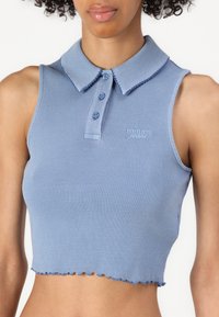 Lys blå ribbet crop top med myk krage, tre knappestolper og flagrende kant. Har en liten brodert logo på brystet.