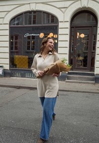 Bež dolg rokav z ovratnikom, združen z zvonastimi modrimi jeans hlačami. Drži šopek, zavit v rjavi papir, hodi po kamniti ulici.