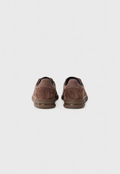 Brown suede sneakers with a rounded shape, cushioned sole, and a contrasting dark rubber outsole. Features embossed logo on the heel.