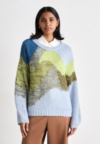 Pull en tricot bleu, vert et marron, avec un motif en vagues. Manches longues, ourlet côtelé, et coupe décontractée. Porté sur une chemise à col.