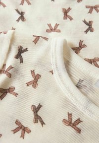 Tissu en coton ivoire arborant un motif répétitif de nœuds marron. La texture est côtelée, avec un col rond et des bords finis.