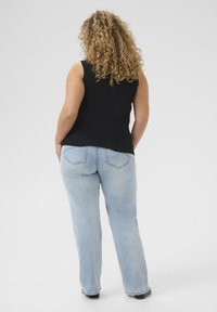 Haut sans manches noir associé à un jean droit bleu clair, avec des poches arrière et une texture douce. Cheveux bouclés partiellement visibles.