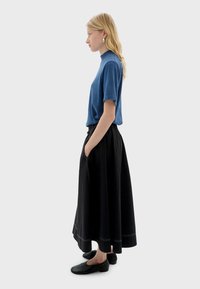 Blauwe top met korte mouwen, zwarte rok tot aan de taille met zakken, en zwarte instapschoenen, allemaal tegen een neutrale grijze achtergrond.