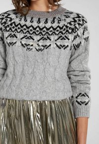 Grå kabelstickad tröja med svartvit mönstrad ok och metalltrådsbroderad halsringning, kombinerad med en glänsande, veckad guldsjal.