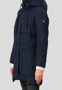Námořnický puffer kabát s kapucí, zipem a knoflíkovým zapínáním, dvěma předními kapsami, elastickými šňůrkami a prošívanou strukturou. Kabát střední délky.