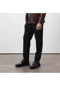 Schwarze Hosen aus einem glatten Stoff, kombiniert mit schwarzen Schnürstiefeln mit einer robusten Sohle. Abgerundet mit einer braunen Lederjacke.