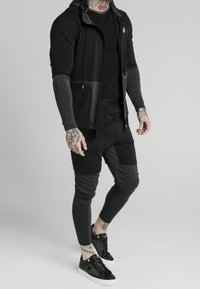 Zwart sportoutfit met een capuchon-jack met rits, een snug t-shirt en taps toelopende broek in verschillende grijstinten; slanke zwarte sneakers.