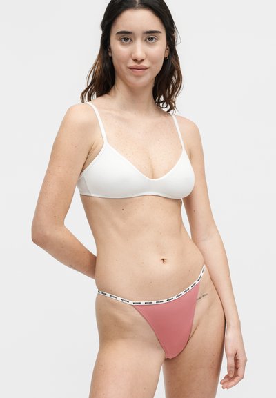 Jeune femme portant un bralette blanc et un string rose avec une ceinture blanche comportant un texte noir, debout devant un fond clair uni.