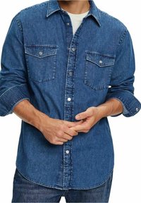 Camisa de mezclilla en azul oscuro con dos bolsillos en el pecho, botón frontal, cuello clásico y puños enrollados. La textura parece resistente y duradera.