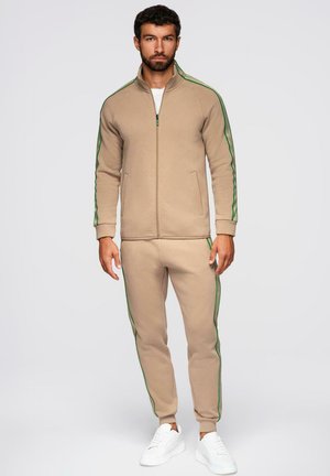 Hombre de pie con chaqueta beige con cremallera y pantalones a juego con rayas verdes, combinados con zapatillas blancas, sobre un fondo liso.
