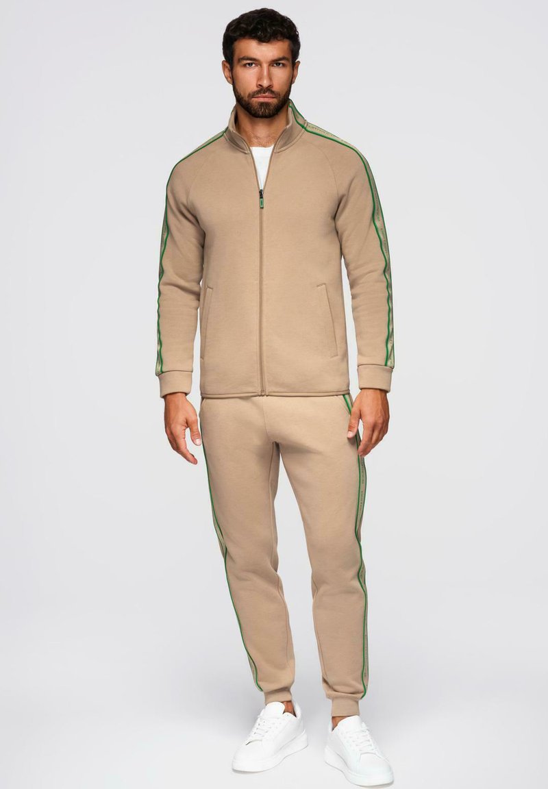 Uomo in piedi che indossa una giacca beige con zip e pantaloni abbinati con strisce verdi, abbinati a scarpe da ginnastica bianche, su sfondo neutro.