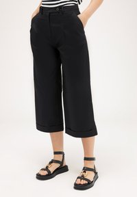 Pantalons noirs coupés, de coupe décontractée, dotés de poches latérales et d'un ourlet à revers. Associés à des sandales noires à lanières avec des accents dorés.