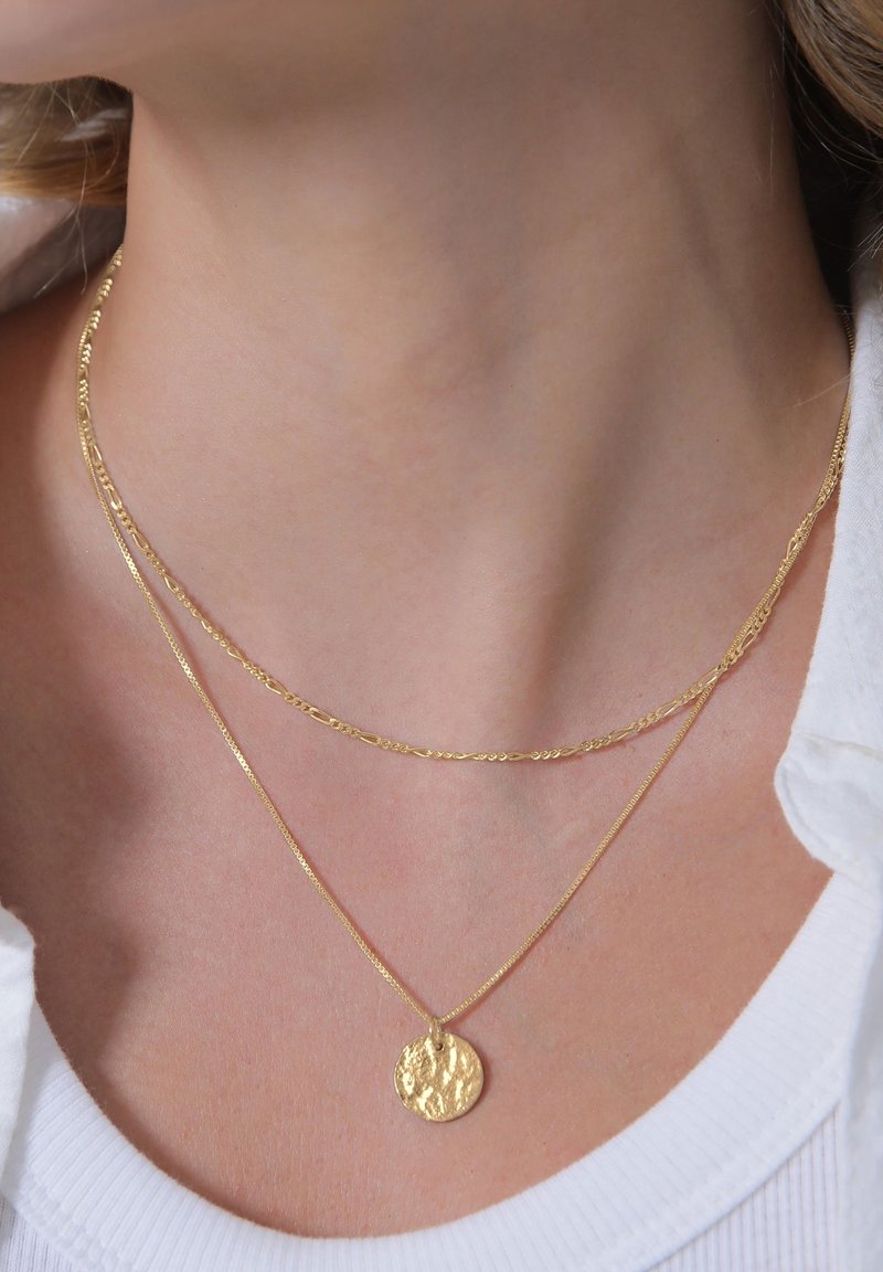 Deux colliers en or délicats autour du cou d'une personne, un chaîne avec un pendentif rond texturé et l'autre une chaîne simple.
