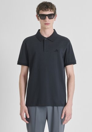 Joven vestido con una camiseta polo negra de manga corta con cuello y puños acanalados, pantalones grises plisados y grandes gafas de sol negras, contra un fondo blanco.