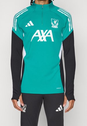 Homme portant un haut d'entraînement Liverpool FC bleu sarcelle et noir avec les logos blancs Adidas et AXA, associé à un pantalon d'entraînement noir avec logos.