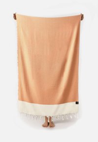 Textieldeken met een perzik-oranje patroon en diamantmotieven, een witte franjerand en een gladde textuur. Handen zichtbaar die de deken vasthouden.