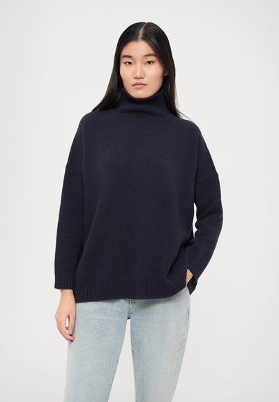 Navy blue turtleneck sweater made of soft knit fabric, featuring a relaxed fit and ribbed cuffs, worn with light blue jeans.