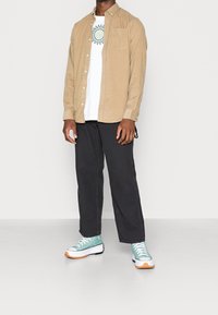 Camisa de pana beige de botones sobre una camiseta gráfica blanca, combinada con pantalones negros y zapatillas en verde azulado con acentos naranjas y suelas blancas.