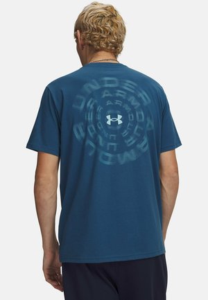 Personne aux cheveux blonds courts portant un t-shirt bleu Under Armour avec un logo circulaire dans le dos.