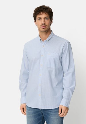 Mand iført lyseblå og hvide stribede skjorte med brystlomme og blå jeans, stående mod en ensfarvet hvid baggrund.