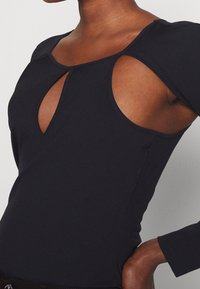 Svart långärmad bodysuit med nyckelhålsutskärning och cut-out detaljer vid axlarna, tillverkad av mjukt och stretchigt material.