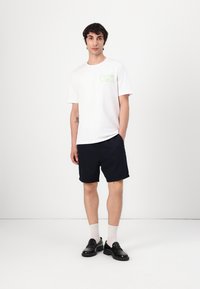 Hvit bomull t-skjorte med grønt grafisk logo, kombinert med marineblå shorts, svarte loafers og lyse sokker. En enkel, avslappet antrekk.