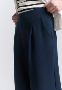 Marineblauwe geplooide broek met een hoge tailleband. Gladde stof met verticale plooien, gecombineerd met een gestreepte top die zichtbaar is bij de taille.